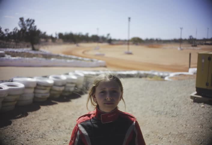The file you uploaded is a very short video or still image showing a young girl wearing a racing suit standing in front of what appears to be a dirt gokart trackshe is looking directly at the camera and there are stacks of white tires and some equ