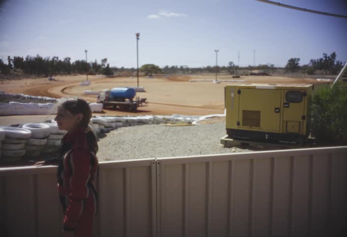 This clip shows a young girl still wearing her racing suit standing next to a white fence with her hand resting on the top railin the background is a dirt race track a large yellow generator and a truck with a blue tank parked near the track