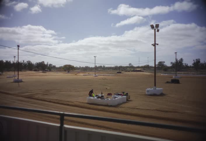 This shot is a wider view of the dirt racing track under a mostly cloudy skytwo people are standing in the centerleft of the frame near a table or platform that has a couple of gokarts and some equipment on it