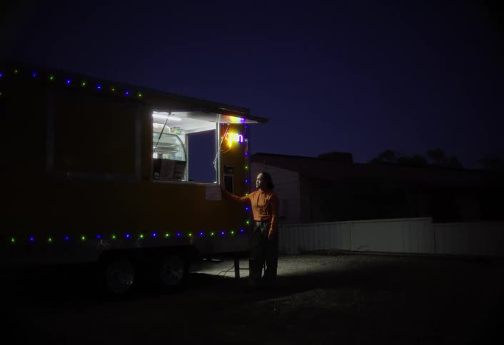 A young woman stands outside a food truck at night purchasing food as the truck glows brightly under its lights