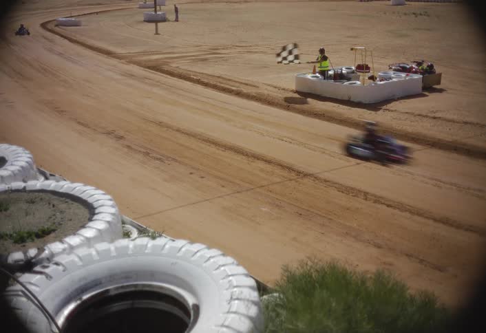 This dynamic shot captures the finish line of the dirt track seen from a slightly elevated position with large white tires in the foregrounda person in a safety vest is waving a checkered flag as a small gokart speeds by on the track