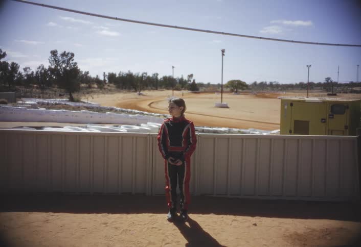 Woman wearing a racing uniform stands near a low wall near a dirt track
