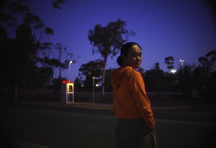 A woman wearing an orange hoodie stands outside at night looking over her shoulder at the camera with a red glowing phone booth visible in the dark background against a deep blue sky