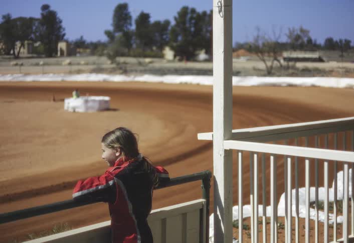 This final clip shows the young girl viewed from the side and back leaning on a white railing as she watches the actionthe dirt track is in the background and several gokarts can be seen driving around the wide curve