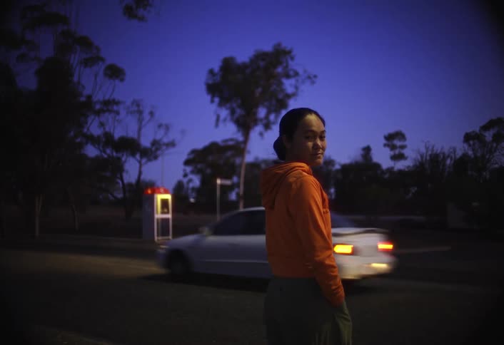 A woman wearing an orange hoodie stands on a dark unlit road at dusk turning to smile at the camera as a car drives past and a phone booth is visible in the background