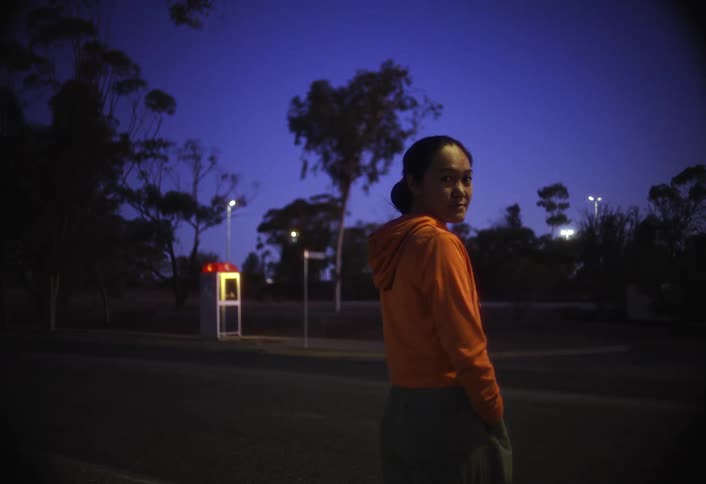 This shot features a woman in an orange hoodie standing outside at dusk or night with a deep blue sky visibleshe turns her head to look directly at the camera and in the background a white and red telephone booth and a truck are faintly v