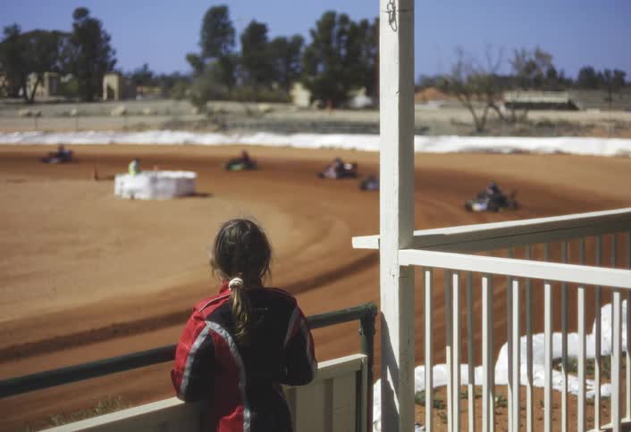 A young racer in a red suit leans on a railing watching karts blur around a dusty oval track