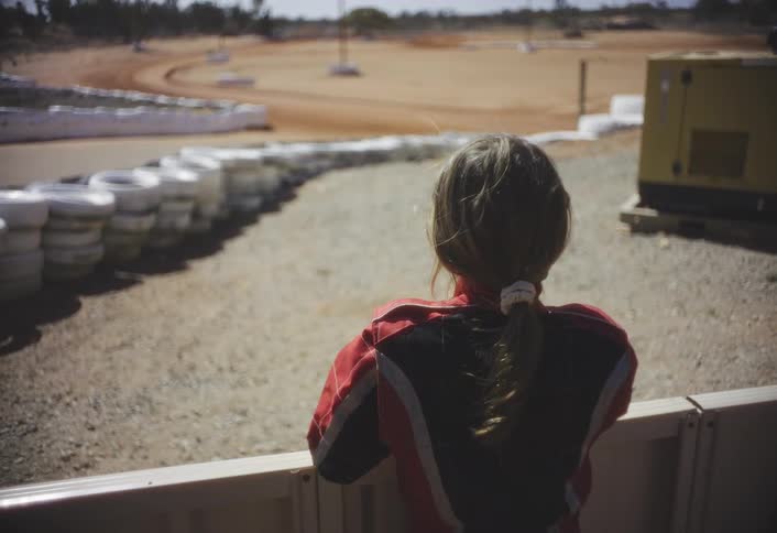 This image is a view from behind the young girl in the racing suit as she leans on a fence looking at the trackthe dirt track curves to the left lined by a long barrier made of stacked white tires with a yellow generator visible on the r