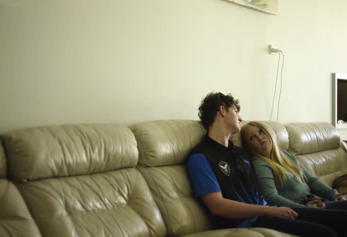 A young man with dark curly hair wearing a blue vest and a young woman with blonde hair are relaxing and talking on a large tan leather couch against a plain white wall