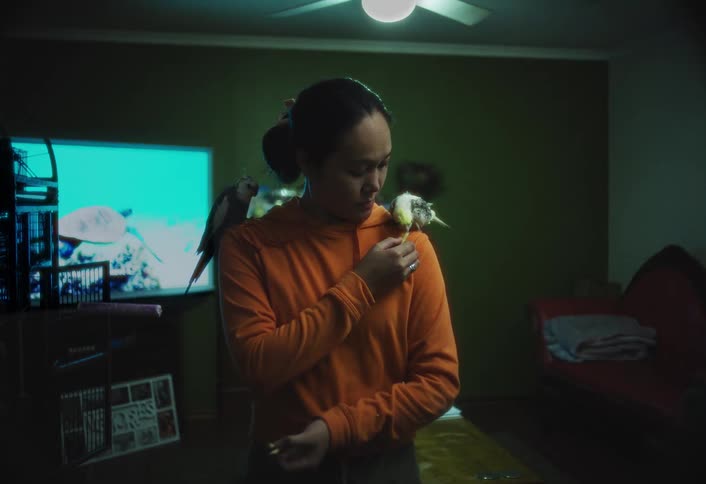 In a dimly lit room illuminated by a television a woman interacts with two pet birds perched on her shoulders