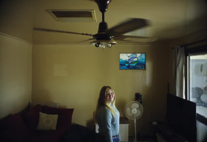 This shot is indoors and features a young woman with long blonde hair standing in a dimly lit living roomshe turns and smiles at the camera with a ceiling fan and a piece of stained glass art visible above her