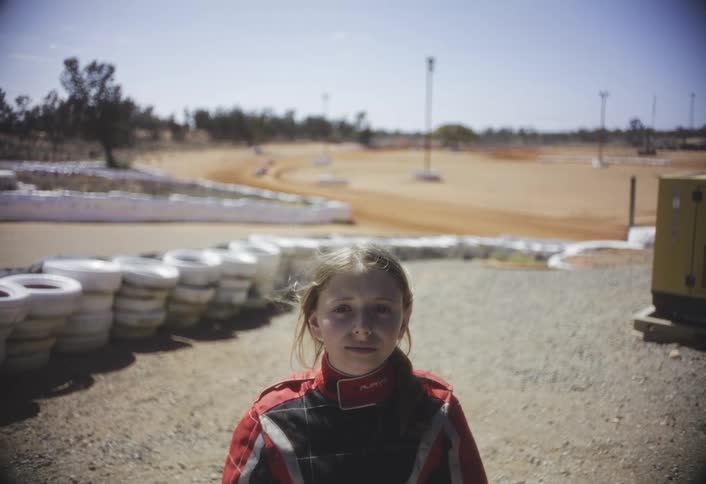 A steady medium shot features a young girl possibly a racer standing outdoors in a red and black racing suit she is looking directly at the camera with a serious expression with a dirt racetrack visible in the background lined by stacked white tires