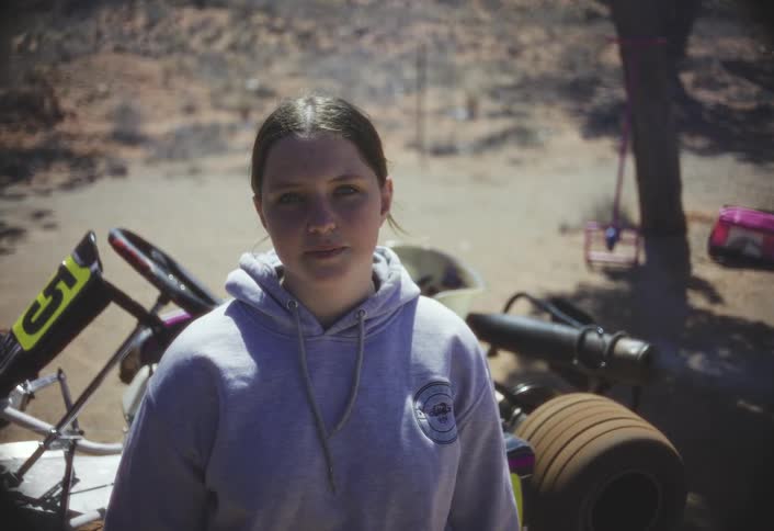 A young woman in a light gray hoodie stands before go kart equipment and stacked tires in a sunlit dusty outdoor area