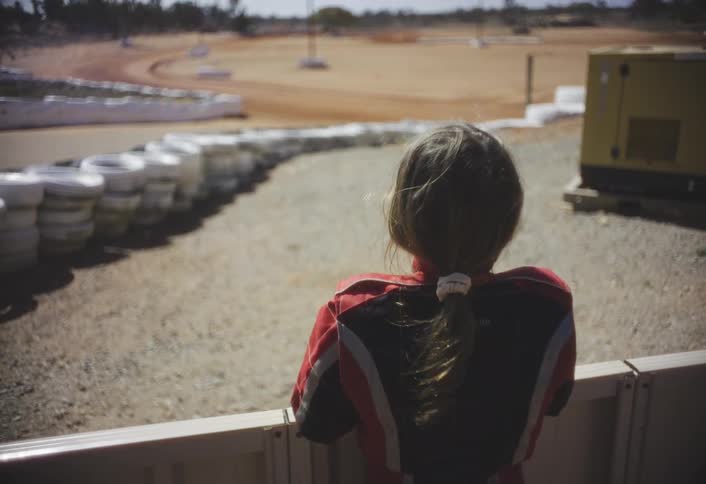 A person in a racing suit looks out over a dirt racetrack under bright daylight