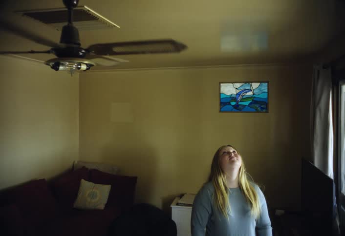 A blonde woman wearing a light blue sweater stands in a dimly lit living room looking up at the ceiling and the moving black ceiling fan with a colorful stained glass artwork visible nearby