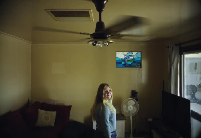 A blonde woman wearing a light blue sweater stands facing the camera in a dimly lit living room next to a black ceiling fan and a floor fan with a small stained glass artwork on the wall