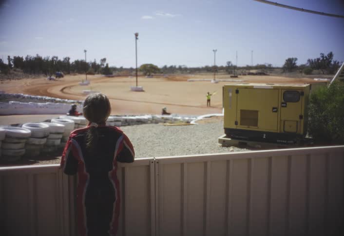 A teenage girl stands behind a fence wearing a sporty jacket watching other kids race by in gokarts on a bright energetic day