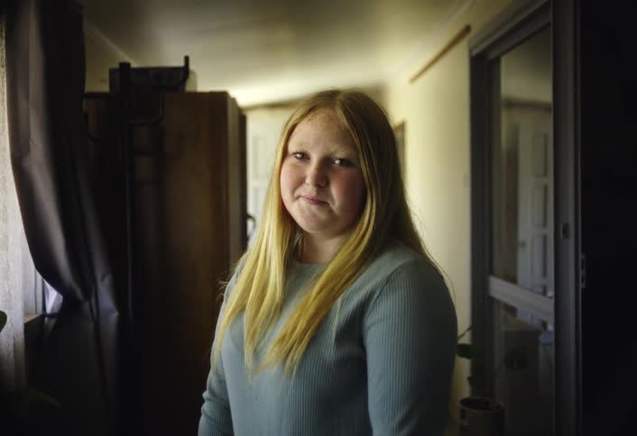 A teenage girl with long blonde hair stands by a window in a narrow hallway giving a shy smile