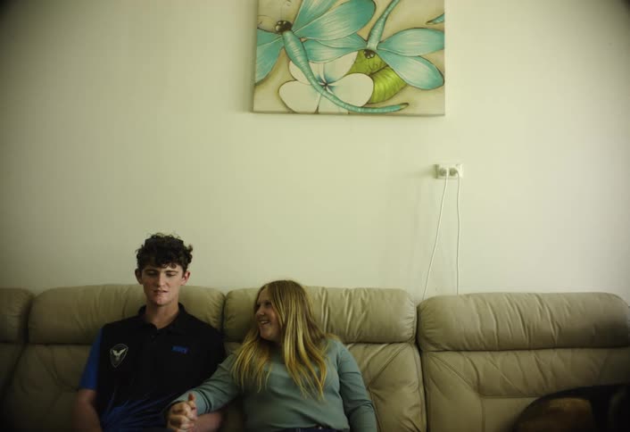 This indoor shot features a young man and a young woman sitting on a beige leatherstyle couchthe man wearing a dark collared shirt is looking forward while the woman with long blonde hair reaches out and takes his hand both smiling sligh