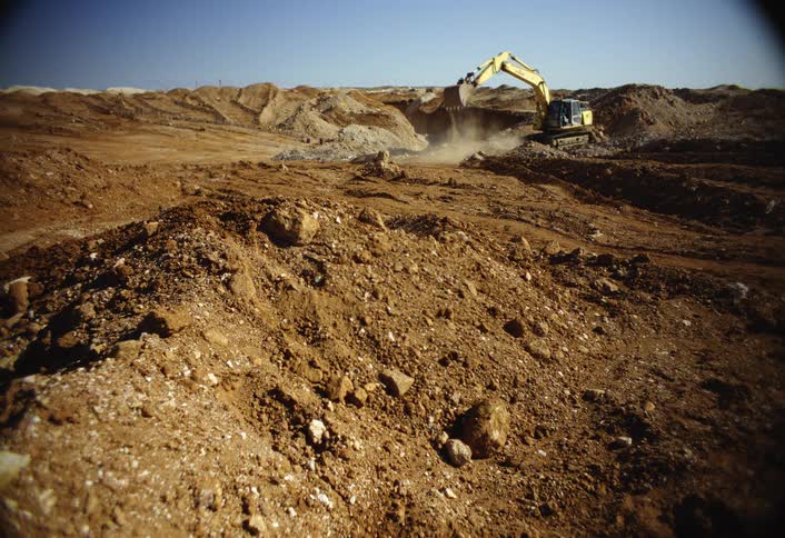 A yellow excavator digs among rocky mounds of brown earth in a sunlit open pit