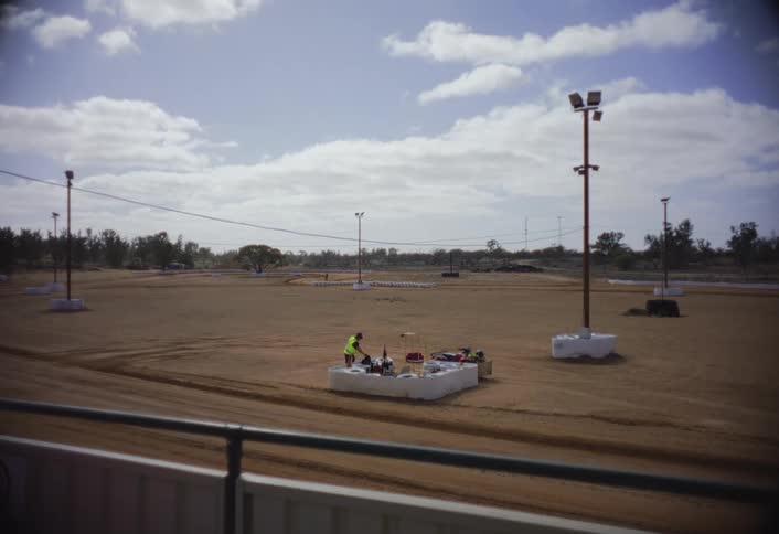 A high wide view of a dirt speedway shows a man wearing a yellow high visibility vest standing at a table next to a starting line surrounded by track lighting poles under a bright cloudy sky