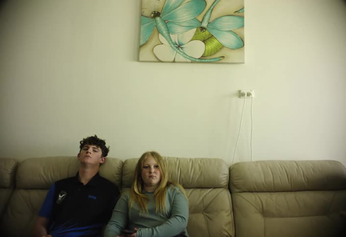 A brother and sister sit quietly on a beige couch in a living room