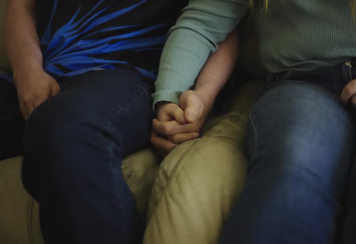 A close view of a man and woman sitting together shows their hands clasped and resting on their legs with the woman wearing a light blue top and the man wearing a blue shirt
