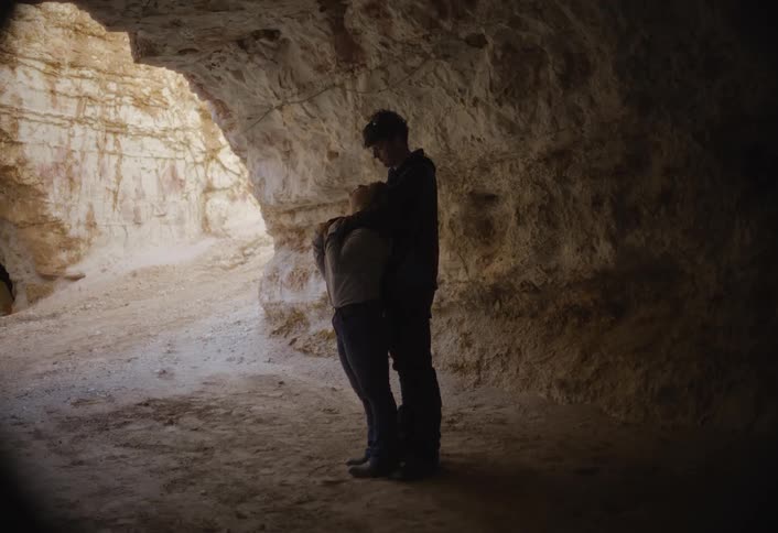 A couple stands embracing inside a dark cave or rock overhang facing the bright light coming from the large opening on the left the light illuminates the sandycolored rock walls at the entrance while casting the two figures in silhouette