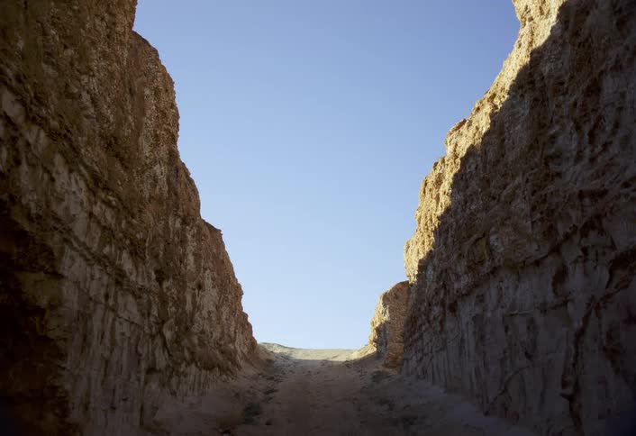 This impressive shot looks up through a narrow canyon or gorge with towering walls of earthen rock on either sidethe natural path opens up to a bright clear blue sky above and a small upward sloping dirt track ahead