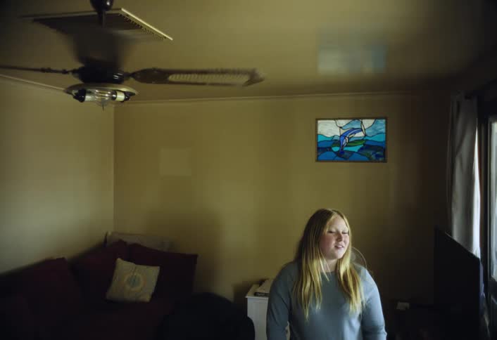 A young blonde woman wearing a light blue sweater stands facing the camera in a dimly lit living room next to a moving black ceiling fan and a colorful stained glass dolphin artwork
