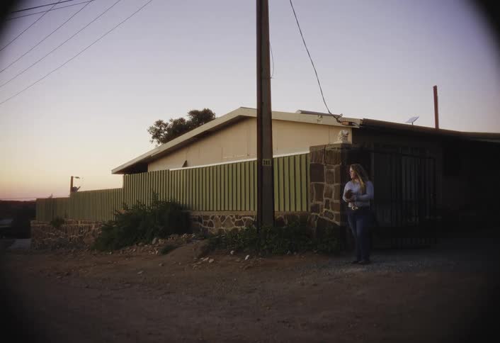 A woman standing outside a house with a stone facade and green fence holding a small object in her hands as she is partially obscured by the shadow of the house and a telephone pole all set against the evening sky over a unpaved area