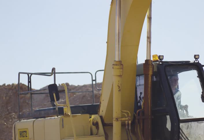 A woman wearing a blue jacket is operating a yellow excavator on a dry dusty construction site looking out from the cab as the machine moves its large arm and bucket