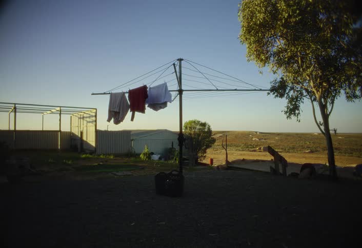 In a rural yard with dark ground a rotating clothesline stands central in the frame holding several pieces of clothing bathed by sunlight backed by various metal sheds and a sunlit tree on the right with an open arid landscape