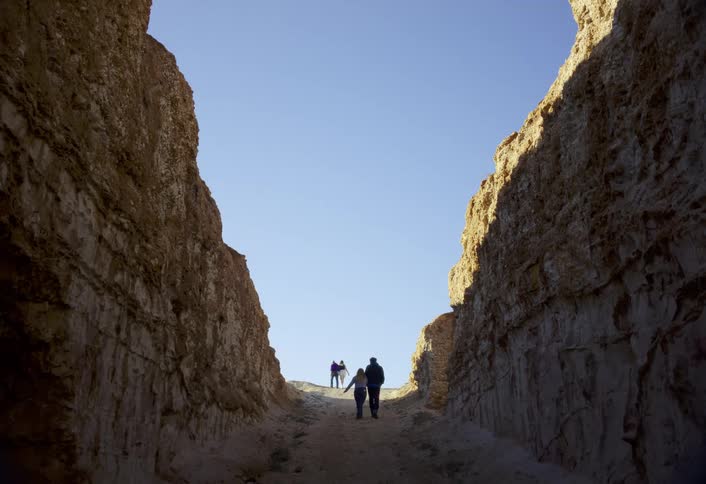 A group of four people walk through a tall rock ravine toward the bright blue sky at the sunlit exit