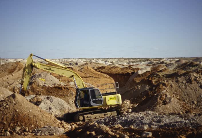 A bright yellow excavator digs into a mound of brown and light colored dirt in a vast sunny open pit mine or quarry under a pale blue sky