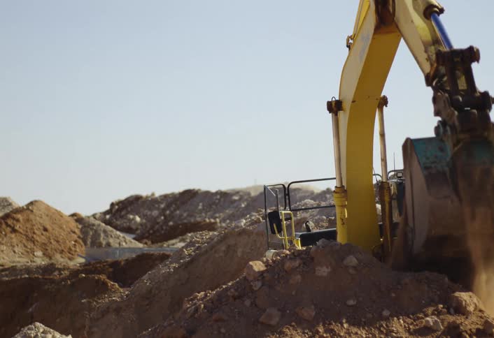 A large bright yellow excavator actively digging in a deep sunlit pit with its articulated arm repeatedly scooping up and moving brown earth amidst surrounding mounds of dirt while the operator is visible inside the cabin performing the work