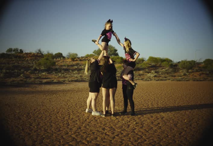 A cheerleading team of five women and one girl in black uniforms performs a cheer stunt pyramid in a dry red dirt field surrounded by scrub brush under a clear blue sky