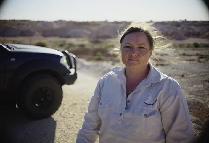 A confident woman wearing a light gray button shirt stands on a dirt road next to a dark truck looking directly at the camera with a dry dusty landscape in the sunlit background