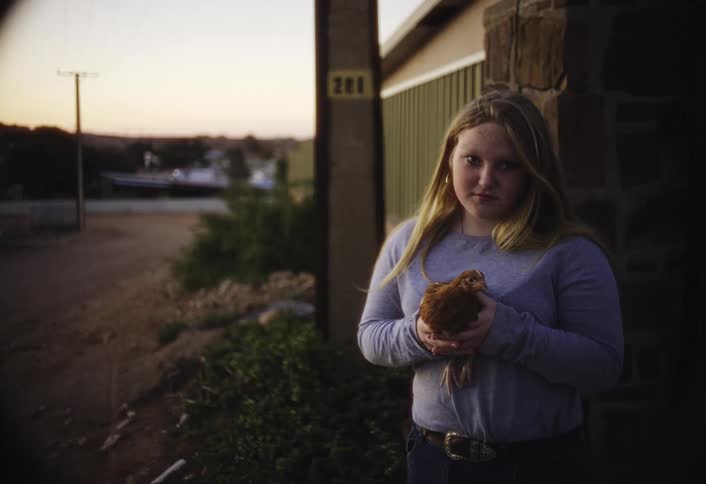 A young blonde woman wearing a gray long sleeve shirt and jeans stands outside a building holding a small brown chicken in her hands and looking thoughtfully at the camera at sunset