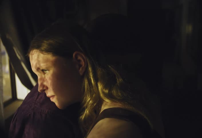 A young woman in dim warm light looks pensively out a window her face half shadowed