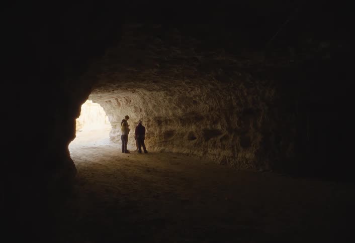 A small group of people stands silhouetted by the light at the far end of a large dark cave or earthen tunnel entrance with rough textured walls