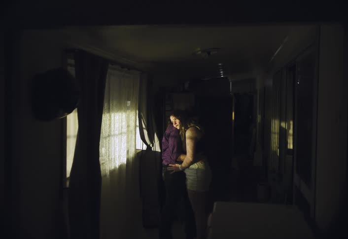 A man and a woman stand in a dim hallway by a window sharing a quiet embrace in a warm beam of light