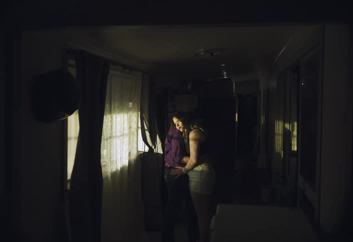 A young man and woman are standing close and embracing in a dark hallway illuminated by a bright shaft of sunlight streaming through a window covered with a dark curtain