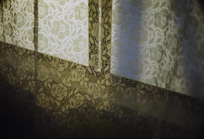 The video is a static closeup shot focusing on a pair of lace curtains drawn across a window through which bright daylight is shiningthe light creates a stark shadow and highlight contrast emphasizing the intricate floral pattern of the sheer fabr