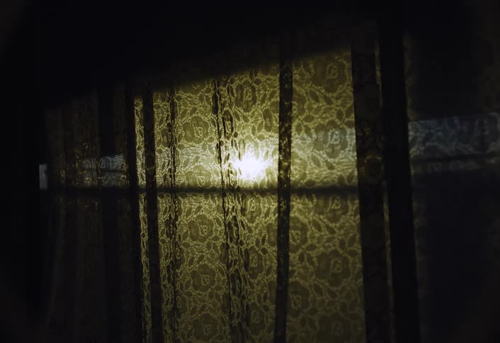 A dimly lit dark interior view shows a bright blurry light source shining through a window covered by a yellow patterned lace curtain and vertical window panes