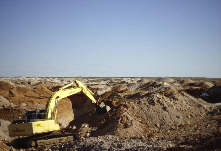 A bright yellow excavator digs into a mound of brown and light colored dirt in a vast dry open pit mine or quarry under a pale blue sky
