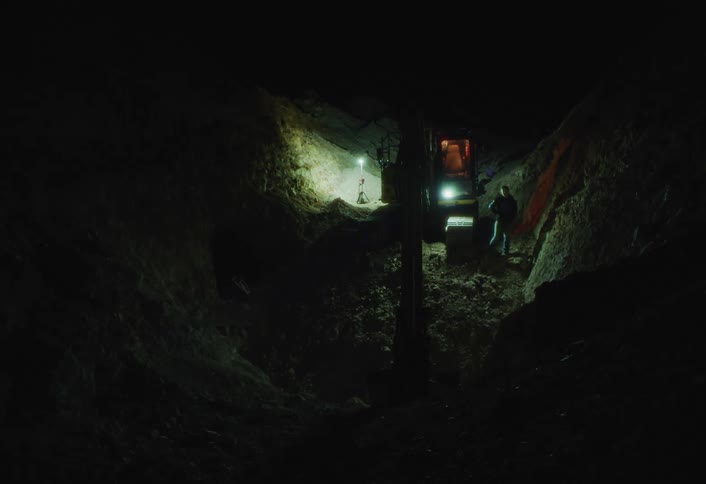 A single bright work light illuminates a person standing next to machinery in an otherwise pitchblack underground tunnel or mine