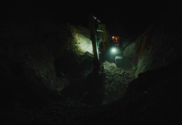 An excavator digs into the rocky ground under dim lights at night