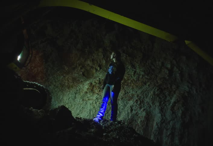 A person stands in a dark cavelike or industrial space illuminated dramatically by a blue light coming from below they are positioned against a rough lightcolored wall with a large yellow beam visible above them