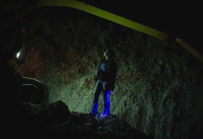 A woman wearing jeans and a dark jacket stands on a pile of dirt in a dark trench using a blue flashlight under a yellow metal beam overhead